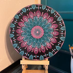 May include: A round, black canvas featuring a detailed mandala design in shades of pink and teal. The mandala is centered with a pink circle and surrounded by intricate patterns. The artwork is displayed on a small wooden easel.