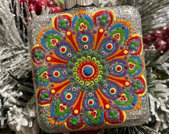 May include: A square, glitter-covered Christmas ornament with a colorful mandala design. The mandala features concentric circles and floral patterns in shades of red, orange, yellow, green, blue, and purple. The ornament is hanging on a snowy, green Christmas tree.