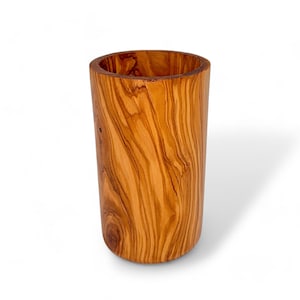 Pode incluir: Um copo ou suporte cilíndrico de madeira de oliveira. O grão natural da madeira é visível em tons de castanho e bege. O copo tem uma parte superior aberta e uma base arredondada, criando um design simples e elegante. Altura aproximada 10 cm.