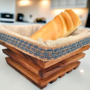 Può includere: Un cestino per il pane in legno con fodera in lino colorata e rifiniture decorative blu. Una pagnotta di pane è all'interno del cestino. Il cestino ha un design unico a strati ed è realizzato in legno naturale.