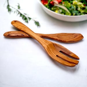 May include: Wooden salad serving set. The set includes a fork and a spoon, both crafted from light brown wood. The fork has three tines, and the spoon has a rounded bowl. The wood grain is visible, adding to the natural aesthetic. A bowl of salad is in the background.