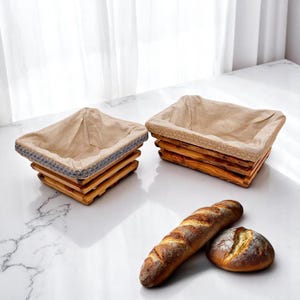 Pode incluir: Duas cestas de pão de madeira com forro de linho sobre uma superfície de mármore, juntamente com uma baguette e um pão redondo. As cestas têm um design rústico com ripas de madeira em camadas e acabamentos decorativos.
