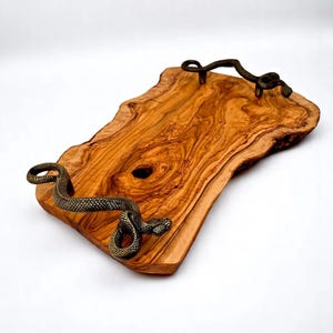 Puede incluir: Bandeja rectangular de madera con veta natural. Tiene asas de serpiente de color bronce. La madera es de color marrón dorado cálido y los bordes tienen forma natural. Aproximadamente 30 cm de largo.