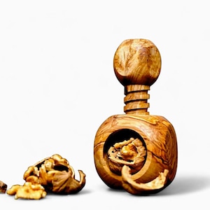 Op de afbeelding: Een houten notenkraker met een bolvormige bovenkant en een schroefdraad. De notenkraker heeft een rond lichaam met een walnoot erin. Een stapel gepelde walnoten staat naast de notenkraker.