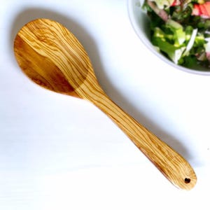 Könnte beinhalten: Ein hellbrauner Holzlöffel mit einem herzförmigen Loch am Ende des Griffs. Der Löffel hat einen breiten, ovalen Kopf und einen langen, schlanken Griff. Die Holzmaserung ist über den gesamten Löffel sichtbar. Ein Küchenutensil.