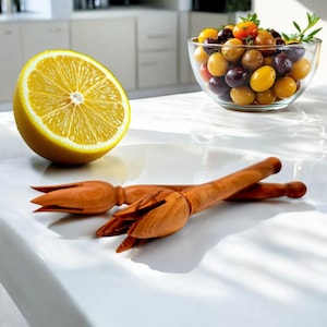 May include: A halved lemon and two wooden citrus reamers are on a white surface. A glass bowl of colourful cherry tomatoes is in the background. The reamers are made of light brown wood.