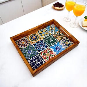 May include: Wooden serving tray with a mosaic tile design. The tray has a rectangular shape with a natural wood frame. The tiles feature a variety of colourful patterns in blue, green, yellow, and red. The tray is on a white surface.