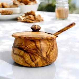 Pode incluir: Uma açucareira de madeira de oliveira com tampa e colher. A tigela apresenta um padrão de grão de madeira natural. A tampa é encimada por um botão decorativo. A colher é colocada na lateral da tigela.