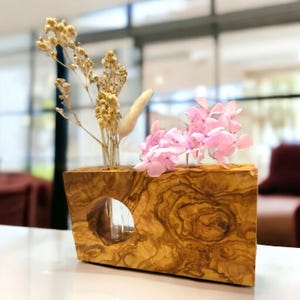 May include: A wooden vase with a natural wood grain finish, holding dried beige flowers and pink hydrangeas. The vase has a cylindrical glass insert and a circular opening on the side. A decorative piece for home decor.