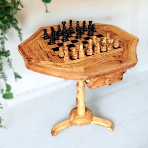 May include: A wooden chess table with a unique octagonal top and a pedestal base. The chess board is inlaid with a black and tan checkered pattern, and the chess pieces are black and tan. The table is made of light brown wood.