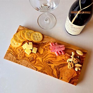 Puede incluir: Una tabla de servir de madera con galletas, cubos de queso y embutidos. La tabla también contiene almendras, anacardos y nueces. Una copa de vino y una botella de vino sugieren una tabla de embutidos.