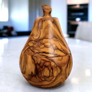 May include: A handcrafted wooden pear-shaped box with a lid. The box is made of olive wood, showcasing a warm, natural color with intricate grain patterns. The lid has a small, curved handle. The box is approximately 6 inches tall.