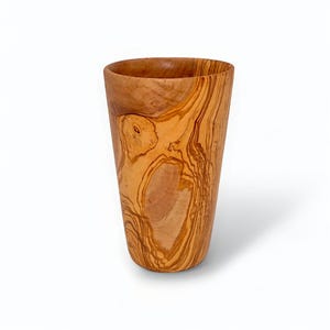 May include: A handcrafted wooden cup with a natural, light brown finish. The cup has a cylindrical shape with a slightly flared top and a smooth, polished surface. The wood grain is visible, adding to its rustic charm.