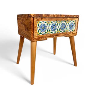 May include: A wooden side table with a drawer featuring a decorative tile design. The table has four tapered legs and is made of light-colored wood. The drawer front showcases a pattern of blue, yellow, and green floral tiles.