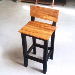 May include: A wooden bar stool with a square seat and backrest. The seat and back are made of light-coloured wood with a natural grain pattern. The frame is black, creating a contrast with the wood. The stool has a simple, modern design.