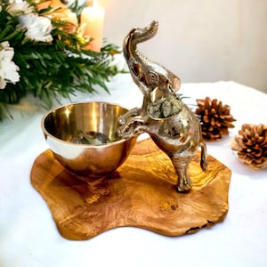 May include: A decorative silver-toned elephant figurine reaching towards a gold-colored bowl. The bowl sits on a wooden, irregularly shaped serving board. The background includes greenery, candles, and pinecones, creating a warm, inviting aesthetic.