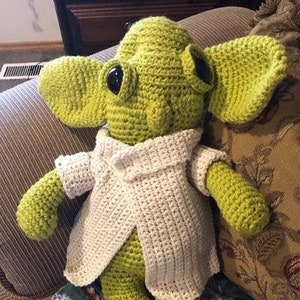 May include: A handmade, crocheted doll resembling a character with large ears. The doll is mainly lime green with black eyes and a cream-coloured jacket. The doll is sitting on a floral patterned cushion.
