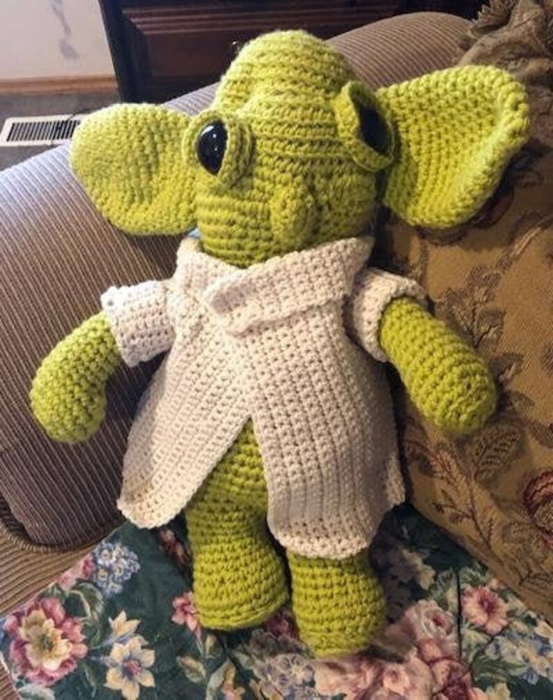 May include: A handmade, crocheted doll resembling a character with large ears. The doll is mainly lime green with black eyes and a cream-coloured jacket. The doll is sitting on a floral patterned cushion.
