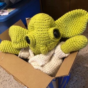 May include: A handmade crochet doll of a character with large ears and black eyes, wearing a white crocheted outfit. The doll is light green and is inside a cardboard box.