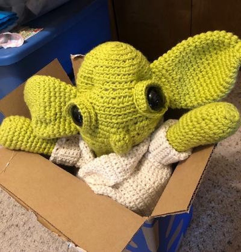 May include: A handmade crochet doll of a character with large ears and black eyes, wearing a white crocheted outfit. The doll is light green and is inside a cardboard box.