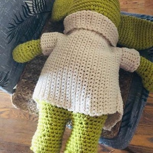 May include: A handmade crochet doll in shades of green and cream. The doll has a pointed hat, long ears, and a cream-coloured top with green arms and legs. The doll is sitting on a chair.