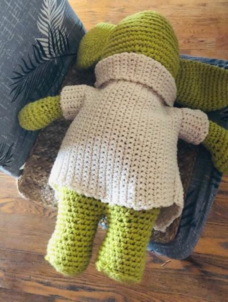 May include: A handmade crochet doll in shades of green and cream. The doll has a pointed hat, long ears, and a cream-coloured top with green arms and legs. The doll is sitting on a chair.