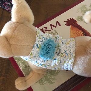 May include: A plush, beige dog toy wearing a white crocheted vest with blue and yellow accents and a blue flower. The dog toy is lying on a decorative sign with a rooster illustration.