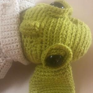 May include: A close-up of a lime green crocheted frog head with large black eyes. The frog has a textured, handmade appearance. A portion of a cream-coloured crocheted item is visible on the left side of the image.