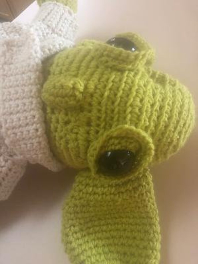 May include: A close-up of a lime green crocheted frog head with large black eyes. The frog has a textured, handmade appearance. A portion of a cream-coloured crocheted item is visible on the left side of the image.