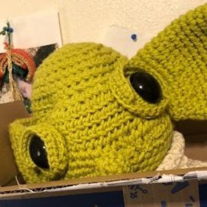 May include: A handmade, lime green crocheted hat with large, black, button eyes and a large ear. The hat is in a cardboard box. The hat is a unique, whimsical design.