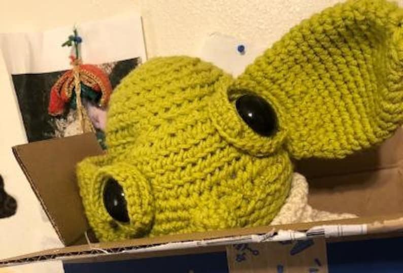 May include: A handmade, lime green crocheted hat with large, black, button eyes and a large ear. The hat is in a cardboard box. The hat is a unique, whimsical design.