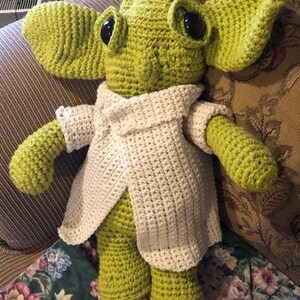 May include: A handmade crochet doll in light green with large ears and black eyes. It is wearing a white crocheted coat. The doll is sitting on floral patterned fabric.
