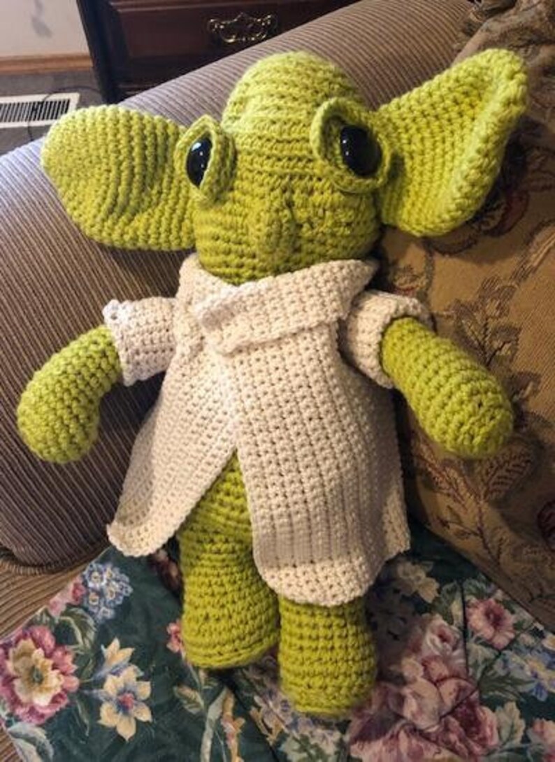 May include: A handmade crochet doll in light green with large ears and black eyes. It is wearing a white crocheted coat. The doll is sitting on floral patterned fabric.