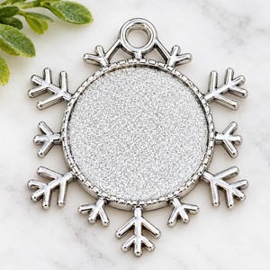May include: A silver-toned metal pendant shaped like a snowflake with a circular center inlay featuring a textured silver glitter finish. The pendant includes a small circular loop at the top for attachment, with six symmetrical arms extending outward.