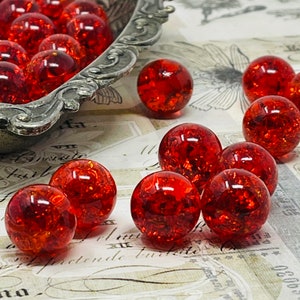 May include: A collection of red glass marbles with a sparkly, iridescent finish. The marbles are scattered on a white surface with a vintage design.