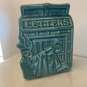 May include: A sage green ceramic mailbox with a glossy finish. The front features the embossed word LETTERS and the letters U.S. above a decorative relief of stacked envelopes. The design includes a rectangular top slot and molded details.