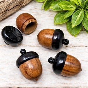 May include: Four acorn-shaped containers crafted from wood, featuring a natural brown nut body and a dark black cap. One container is shown open, revealing a hollow interior, while the other three are closed and include a small loop at the top.