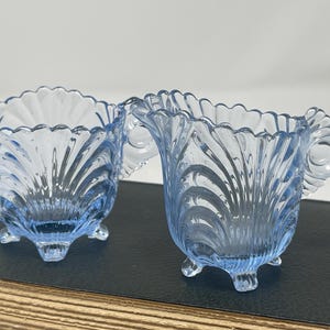 May include: A light blue glass sugar bowl and creamer set featuring a scalloped rim, a ribbed shell-like pattern, and three small feet. The sugar bowl has two side handles, while the creamer features a pouring spout and a single side handle.