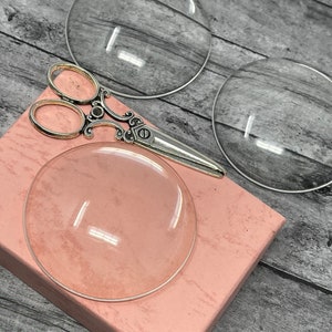 May include: Two clear glass dome shapes, one on a pink box and one on a wooden surface, with a pair of silver scissors in between.