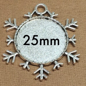 May include: Silver-colored snowflake-shaped pendant with a 25mm round inset. The snowflake design features six symmetrical arms with detailed tips. The pendant has a loop at the top for hanging. The text "25mm" is printed in black.