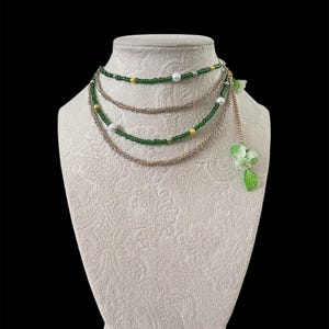 Puede incluir: Collar de varias hebras exhibido en un maniqu&iacute; blanco. El collar presenta cuentas verdes, cadena dorada y detalles de perlas blancas. Un racimo de flores y hojas verdes cuelga del lateral. El fondo es negro.