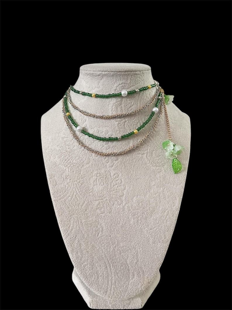 Puede incluir: Collar de varias hebras exhibido en un maniqu&iacute; blanco. El collar presenta cuentas verdes, cadena dorada y detalles de perlas blancas. Un racimo de flores y hojas verdes cuelga del lateral. El fondo es negro.