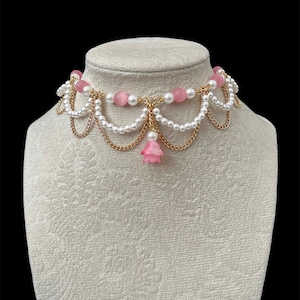 May include: A decorative choker necklace with white pearls, pink beads, and gold-tone chain details. A small pink flower pendant hangs from the centre. The necklace is displayed on a mannequin against a black backdrop.