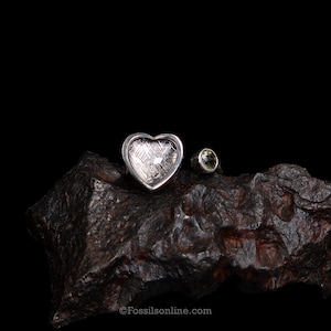 May include: A silver ring featuring a heart-shaped inlay with a cross-hatched Widmanstätten pattern and a small, round, olive-green gemstone set in a bezel. The ring is positioned on a dark, textured, metallic-looking rock fragment.