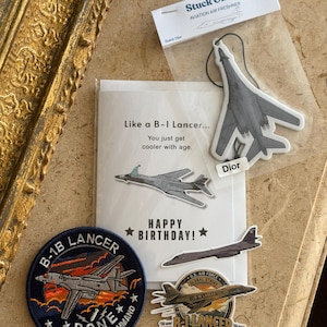 May include: A collection of B-1 Lancer themed items including a greeting card with the text Like a B-1 Lancer... You just get cooler with age. HAPPY BIRTHDAY!, a grey airplane air freshener, a circular embroidered patch, and a die-cut sticker.
