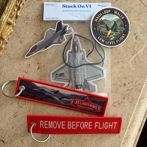 May include: A collection of aviation-themed items including an F-35 air freshener in a clear package labeled Stuck On V1, a circular Mach Loop Wales sticker, a die-cut F-35 sticker, and two red fabric keychains with metal rings.