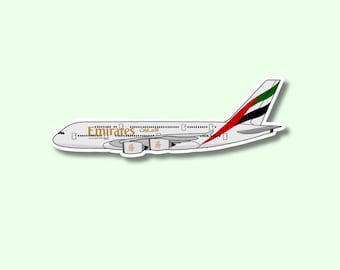 Puede incluir: Una pegatina de un avión Emirates blanco con letras doradas. La aleta de cola presenta los colores de la bandera de los Emiratos Árabes Unidos: rojo, negro, verde y blanco. La pegatina está sobre un fondo verde claro.