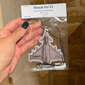Puede incluir: Un ambientador gris con forma de Eurofighter Typhoon en un paquete de plástico transparente. La tarjeta superior dice Stuck On V1 AVIATION AIR FRESHNER y Scent: Oud. El ambientador presenta una ilustración detallada de un avión de combate.