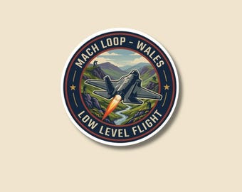 May include: A circular sticker with a jet plane graphic. The sticker reads "MACH LOOP - WALES LOW LEVEL FLIGHT" in a navy blue border. The jet plane is gray with an orange flame, flying over a green landscape.