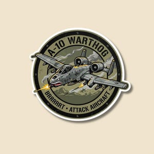 Könnte beinhalten: A-10 Warthog-Angriffsflugzeug-Aufkleber. Der Aufkleber zeigt eine detaillierte Illustration des Flugzeugs mit dem Text "A-10 WARTHOG ATTACK AIRCRAFT" in einem kreisförmigen Design. Das Flugzeug feuert mit einer Berglandschaft im Hintergrund.